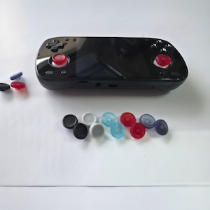 May include: A black handheld gaming console with a glossy screen, a directional pad, and action buttons labeled X, Y, A, and B. Two red joystick caps are attached to the device, with additional loose joystick caps in black, white, blue, and red.