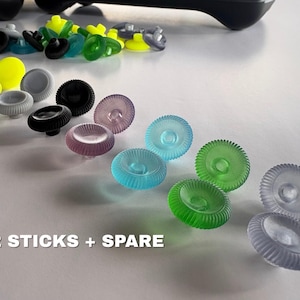 May include: A collection of circular, ridged controller thumbsticks in various translucent and solid colors including black, grey, green, blue, yellow, and purple. The text 2 STICKS + SPARE is printed in white at the bottom left of the image.