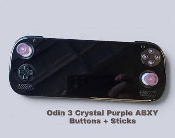 May include: A black handheld gaming console featuring crystal purple analogue sticks and ABXY buttons. The device has a glossy black screen area, a directional pad, and two speaker grilles. Text below reads: Odin 3 Crystal Purple ABXY Buttons + Sticks.