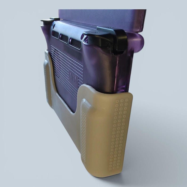 Puede incluir: Una consola de juegos port&aacute;til con carcasa morada transl&uacute;cida y botones superiores negros, asegurada en una funda de agarre beige texturizada. La consola presenta una rejilla de ventilaci&oacute;n trasera y la marca AYN grabada en el panel posterior.