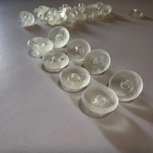 May include: A collection of round, clear, translucent buttons featuring a textured, ribbed edge and a central shank attachment point. These small, circular fasteners are arranged in a loose cluster, displaying a glossy finish and a distinct pattern.
