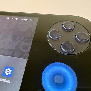 May include: Close-up of a black handheld gaming device. The device features a directional pad with four black buttons and a blue joystick. The screen displays icons and the word "Settings". The battery indicator shows 90% charge.