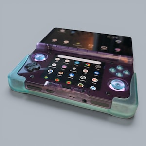Puede incluir: Una consola de juegos port&aacute;til con carcasa morada transl&uacute;cida y protector de silicona azul claro. El dispositivo cuenta con dise&ntilde;o de doble pantalla, cruceta, dos joysticks anal&oacute;gicos y cuatro botones de acci&oacute;n que muestran una interfaz digital.