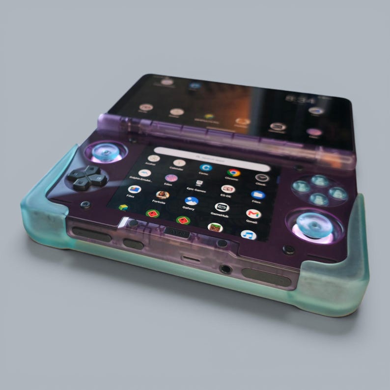 Puede incluir: Una consola de juegos port&aacute;til con carcasa morada transl&uacute;cida y protector de silicona azul claro. El dispositivo cuenta con dise&ntilde;o de doble pantalla, cruceta, dos joysticks anal&oacute;gicos y cuatro botones de acci&oacute;n que muestran una interfaz digital.
