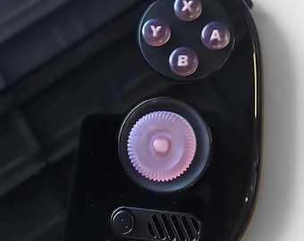 May include: A black handheld gaming console featuring a glossy finish, a circular light purple textured joystick, and four translucent purple action buttons labelled with the letters X, Y, A, and B. A small speaker grille and a button are visible below.