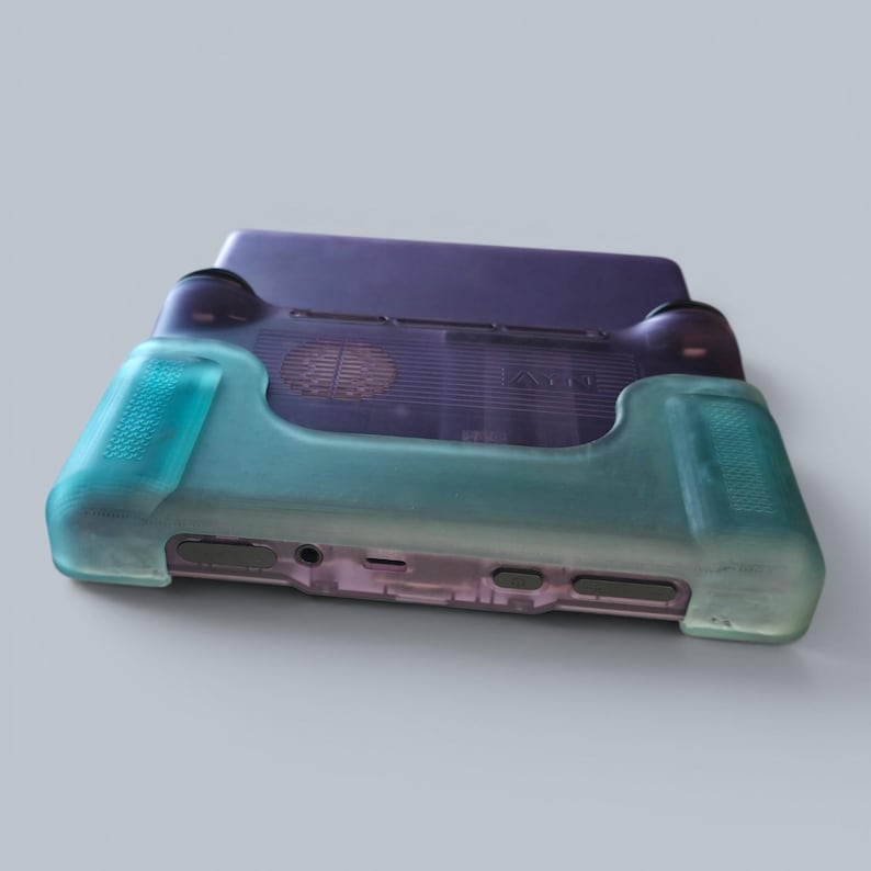 Puede incluir: Una consola de juegos port&aacute;til con una carcasa p&uacute;rpura transl&uacute;cida y una funda protectora de silicona en un degradado de color verde azulado. El dispositivo incluye una rejilla de ventilaci&oacute;n circular, varios puertos y botones inferiores.