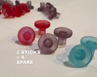 May include: A collection of small, translucent, circular knobs with ridged edges in red, purple, and blue. The knobs feature a central cylindrical stem. The text 2 STICKS + A SPARE is printed in white on the left side of the image.