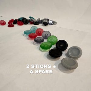 May include: A collection of small, circular, ridged plastic components in various colors including red, green, black, and gray. The items are arranged in rows and scattered piles. The text 2 STICKS + A SPARE is printed in white on the lower portion.