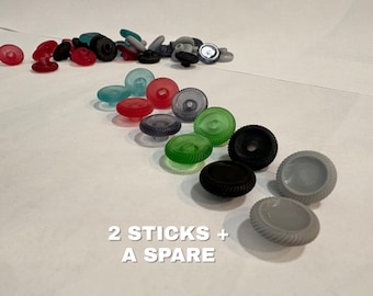 May include: A collection of small, circular, ridged plastic components in various colors including red, green, black, and gray. The items are arranged in rows and scattered piles. The text 2 STICKS + A SPARE is printed in white on the lower portion.