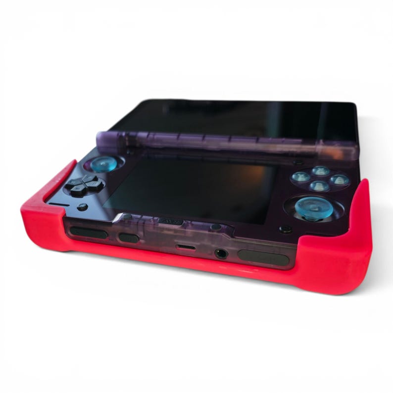 Puede incluir: Una consola de juegos port&aacute;til con una carcasa de pl&aacute;stico morado transl&uacute;cido y una funda protectora de silicona de color rojo brillante. El dispositivo incluye una pantalla negra, un pad direccional, cuatro botones de acci&oacute;n y dos joysticks azules.