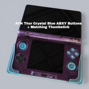 May include: A purple handheld gaming console featuring AYN Thor Crystal Blue ABXY buttons and matching thumbsticks. The device has a dual-screen layout with a black display and a translucent purple shell. Text reads: AYN Thor Crystal Blue ABXY Buttons + Matching Thumbstick.