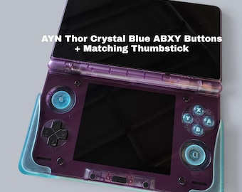 May include: A purple handheld gaming console featuring AYN Thor Crystal Blue ABXY buttons and matching thumbsticks. The device has a dual-screen layout with a black display and a translucent purple shell. Text reads: AYN Thor Crystal Blue ABXY Buttons + Matching Thumbstick.