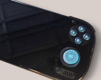May include: A black handheld gaming console featuring a glossy screen, a light blue circular joystick, and four translucent light blue action buttons labelled with the letters A, B, X, and Y. A small power button and a speaker vent are visible below.