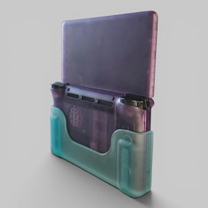 Puede incluir: Una consola de juegos port&aacute;til con una carcasa morada transl&uacute;cida, insertada en una funda protectora de color verde azulado semitransparente. El dispositivo muestra una vista trasera con ranuras de ventilaci&oacute;n, botones y un agarre lateral texturizado.
