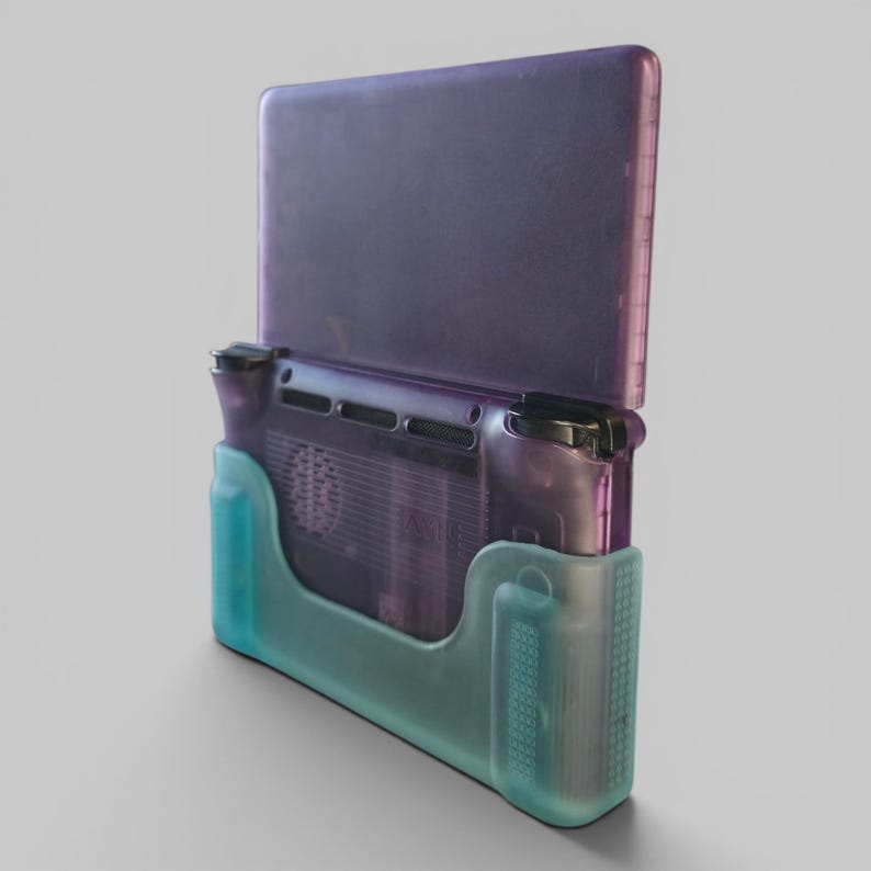 Puede incluir: Una consola de juegos port&aacute;til con una carcasa morada transl&uacute;cida, insertada en una funda protectora de color verde azulado semitransparente. El dispositivo muestra una vista trasera con ranuras de ventilaci&oacute;n, botones y un agarre lateral texturizado.