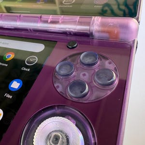 May include: Close-up of a purple handheld gaming device. The device features a screen displaying app icons, a directional pad, and four circular buttons. The device has a glossy finish and a transparent hinge.