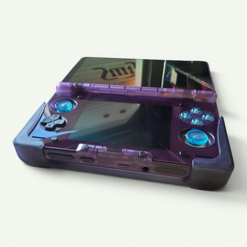 Puede incluir: Una consola de juegos port&aacute;til con carcasa morada transl&uacute;cida, que presenta una pantalla negra, una cruceta, cuatro botones de acci&oacute;n azul claro y dos joysticks anal&oacute;gicos azul claro. El dispositivo est&aacute; dentro de un marco protector morado.