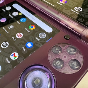 May include: A close-up of a purple handheld gaming device with two screens. The left screen displays app icons, including Gmail, Chrome, and Gallery. The right screen shows game icons. The device has a circular button layout with A, B, X, and Y buttons.