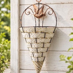 Puede incluir: Una maceta de pared en forma de cono con una cesta de cuerda tejida en color blanco roto, sostenida por un marco de metal de color bronce oscuro. La parte superior del marco incluye una figura decorativa de colibrí y un aro para colgar.