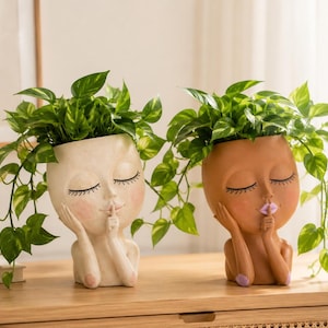 Peut inclure: Deux pots de fleurs en forme de tête avec les yeux fermés et un doigt sur les lèvres dans un geste de silence. L'un est blanc cassé avec des joues roses, l'autre est marron avec des lèvres violettes. Ils contiennent des plantes pothos.