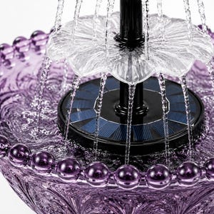 May include: A purple glass water fountain featuring a beaded rim and intricate embossed patterns. The fountain has a central black solar panel base with two clear, tiered, flower-shaped tiers that distribute water in a cascading circular motion.