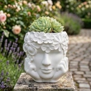 Peut inclure: Un pot de fleurs en céramique blanche en forme de tête humaine avec les mains posées sous le menton. La tête est ornée d'une couronne de fleurs sculptées. Une plante succulente verte est plantée au sommet, émergeant de la couronne florale.