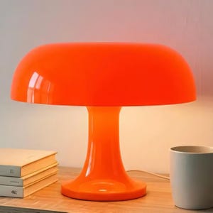 Peut inclure: Une lampe de table orange vif en forme de champignon. La lampe a un abat-jour arrondi et une base en piédestal. La lampe est en matériau brillant et repose sur une surface en bois. Des livres et une tasse blanche sont également visibles.