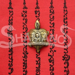 Può includere: Un tessuto rosso con scritte nere e un amuleto a forma di tartaruga in bronzo. L'amuleto presenta una figura seduta all'interno del guscio della tartaruga. La parte superiore del tessuto presenta testo in lingua straniera.