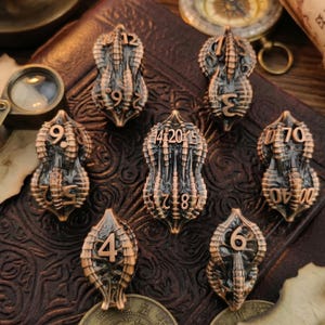 Puede incluir: Una colección de siete dados en forma de caballito de mar, de color bronce, con numeración blanca. Los dados están dispuestos sobre una superficie de cuero marrón oscuro, con una lupa y una brújula en el fondo. Dados para juegos de mesa.