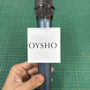 May include: A handheld microphone with a dark grey mesh head and a dark grey body. A white rectangular sign with the word "OYSHO" in black capital letters is attached to the microphone.