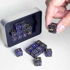 Puede incluir: Un juego de dados de seis caras de color azul oscuro con grabados dorados de cabezas de lobo y símbolos rúnicos. Los dados se guardan en una caja metálica rectangular de color plateado con un inserto de espuma negra en su interior.