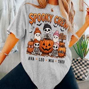May include: A light gray t-shirt featuring a graphic with the text "SPOOKY CREW" in orange. The design includes skeletons and ghosts wearing colorful beanies, pumpkins, bats, and a "PUMPKIN PATCH" sign. The names "AVA", "LEO", "MIA", and "THEO" are printed below.