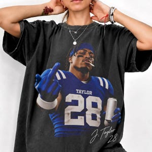 Pode incluir: Uma t-shirt cinzento-carvão de manga curta com uma impressão gráfica de um jogador de futebol americano com uma camisola azul com o nome TAYLOR e o número 28. O jogador usa uma fita azul, segura uma lata prateada e faz um gesto com a mão.