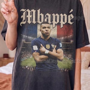 Może przedstawiać: Ciemnoszara koszulka z grafiką Kyliana Mbapp&eacute; w niebieskiej koszulce, na tle francuskiej flagi. Słowo "Mbapp&eacute;" jest wydrukowane w stylu vintage. Koszulka ma postarzany, vintage wygląd.