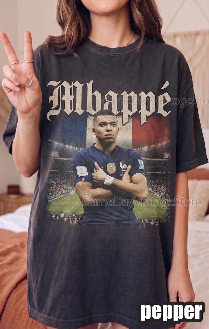 Może przedstawiać: Ciemnoszara koszulka z grafiką Kyliana Mbapp&eacute; w niebieskiej koszulce, na tle francuskiej flagi. Słowo "Mbapp&eacute;" jest wydrukowane w stylu vintage. Koszulka ma postarzany, vintage wygląd.
