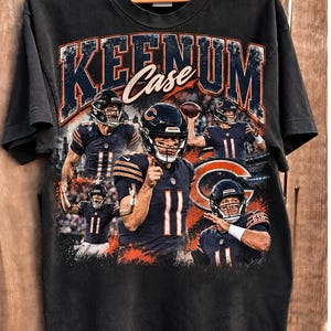 Può includere: Una t-shirt nera a maniche corte con una stampa grafica di un giocatore di football in divisa blu navy con il numero 11. Il testo KEENUM Case è stampato in alto a grandi lettere, circondato da diverse immagini d'azione dell'atleta.