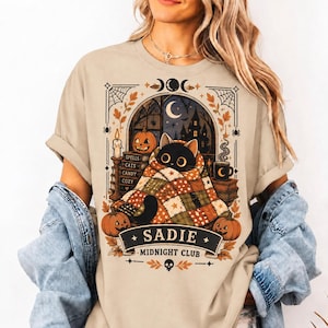 Puede incluir: Una camiseta beige con un gráfico de un gato negro envuelto en una manta de retazos. El diseño incluye calabazas, libros etiquetados como Spells, Cats, Candy, Cozy, una luna creciente y una pancarta con el texto SADIE MIDNIGHT CLUB.