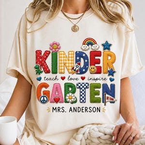May include: A cream-coloured t-shirt featuring the word KINDERGARTEN in colourful, patterned block letters. The design includes icons like a rainbow, rocket, smiley face, and flowers. Below, the text reads teach love inspire and MRS. ANDERSON.