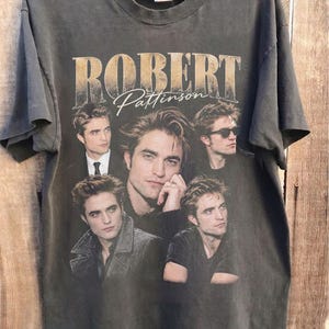 May include: A charcoal grey short-sleeve t-shirt featuring a collage of five photographic portraits of Robert Pattinson. The text "ROBERT Pattinson" is printed in a large, distressed beige font across the top of the graphic design.