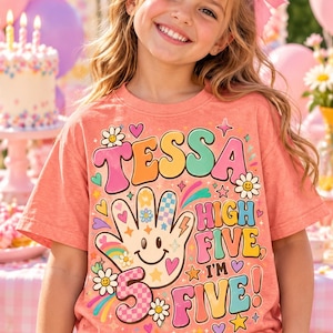 May include: A coral-colored t-shirt featuring a graphic print with the text TESSA HIGH FIVE I'M 5 FIVE. The design includes a smiling hand icon, flowers, stars, hearts, and rainbow elements in pastel shades of pink, yellow, blue, and orange.
