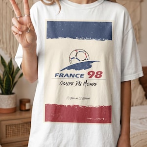 May include: A white t-shirt featuring a graphic print with a football, the text FRANCE 98, COUPE DU MONDE, and 10 JUIN AU 12 JUILLET. The design includes a blue horizontal bar at the top and a red horizontal bar at the bottom with a distressed texture.