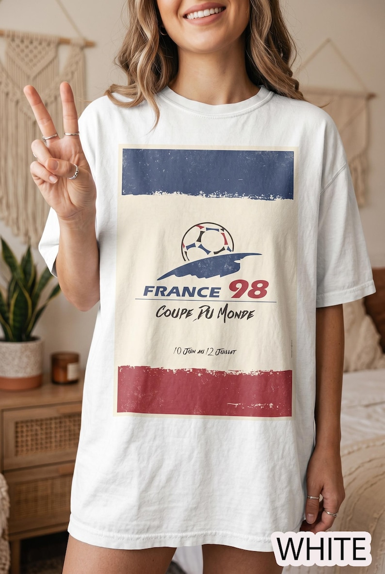 May include: A white t-shirt featuring a graphic print with a football, the text FRANCE 98, COUPE DU MONDE, and 10 JUIN AU 12 JUILLET. The design includes a blue horizontal bar at the top and a red horizontal bar at the bottom with a distressed texture.