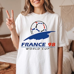 May include: A white short-sleeve t-shirt featuring a graphic of a soccer ball above a blue abstract shape. Below the graphic, the text reads FRANCE 98 in blue and red, followed by the word WORLD CUP in black capital letters on the chest area.