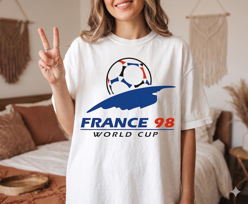 May include: A white short-sleeve t-shirt featuring a graphic of a soccer ball above a blue abstract shape. Below the graphic, the text reads FRANCE 98 in blue and red, followed by the word WORLD CUP in black capital letters on the chest area.
