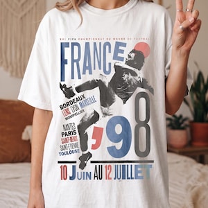 K&ouml;nnte beinhalten: Ein wei&szlig;es T-Shirt mit einem Grafikdruck f&uuml;r die FIFA Fu&szlig;ball-Weltmeisterschaft 1998 in Frankreich. Das Design enth&auml;lt den Text "FRANCE 98", "10 JUIN AU 12 JUILLET" sowie eine Liste franz&ouml;sischer St&auml;dte wie Bordeaux, Lens, Lyon, Marseille und Paris.