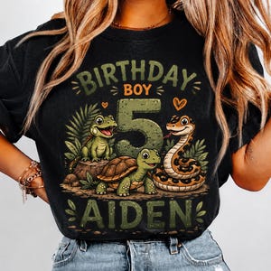 May include: A black t-shirt featuring a graphic print with the text "BIRTHDAY BOY" and "AIDEN" surrounding a large number "5". The design includes illustrations of a green lizard, a turtle, and a snake amidst tropical leaves and small orange hearts.
