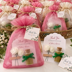 May include: A pink organza drawstring favor bag containing a card that reads Thank You For being a part of my special night Kubra's Henna, two white artificial roses, and a small glass bottle filled with green powder and a green ribbon.