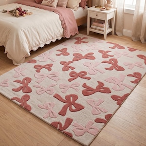 Può includere: Un tappeto rettangolare caratterizzato da un motivo ripetuto di fiocchi in varie tonalità di rosa e rosa antico su uno sfondo beige chiaro. I fiocchi sono distribuiti sulla superficie con diverse orientazioni, creando un effetto visivo strutturato.