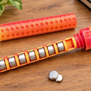 May include: A cylindrical orange and yellow gradient battery storage case with a removable cap. The case features a grid of circular cutouts and holds eight silver button cell batteries in a row. Two additional button cell batteries lie nearby.