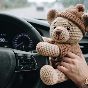 May include: A light brown crocheted teddy bear wearing a matching dark brown knit hat. The bear has dark plastic eyes, a stitched nose, and a textured yarn body. It is positioned near a black steering wheel with a silver Hyundai logo in the center.
